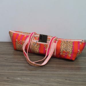 May include: A long, cylindrical fabric bag featuring a vibrant orange and pink striped pattern with repeating tiger illustrations. The bag includes two matching striped carrying straps and a zippered top opening with a black smartphone tucked inside.