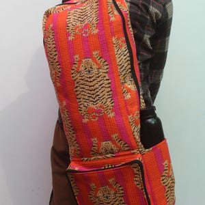 May include: A rectangular backpack featuring a vibrant orange and pink striped pattern with a repeating tiger print. The bag includes a large main compartment, a front zippered pocket, and a side pocket containing a black water bottle.