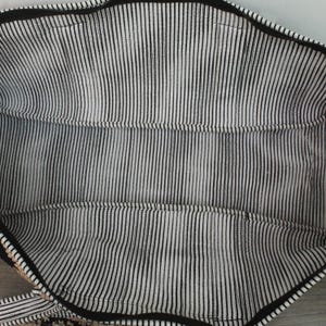 May include: The interior of a tote bag featuring a black and white vertical pinstripe fabric lining. The bag has a black zipper closure along the top edge and visible stitched seams along the sides and bottom, creating a structured rectangular compartment.