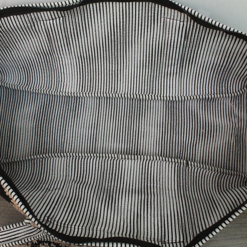 May include: The interior of a tote bag featuring a black and white vertical pinstripe fabric lining. The bag has a black zipper closure along the top edge and visible stitched seams along the sides and bottom, creating a structured rectangular compartment.