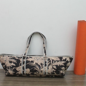 May include: A long, rectangular black and beige floral patterned bag with two matching handles and a top zipper closure. The bag features a detailed botanical print and is positioned next to a tall, solid orange cylindrical yoga mat.
