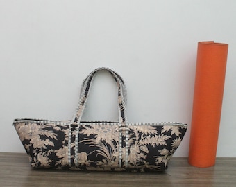 May include: A long, rectangular black and beige floral patterned bag with two matching handles and a top zip closure. The bag features a detailed botanical print and is positioned next to a tall, solid orange cylindrical yoga mat.