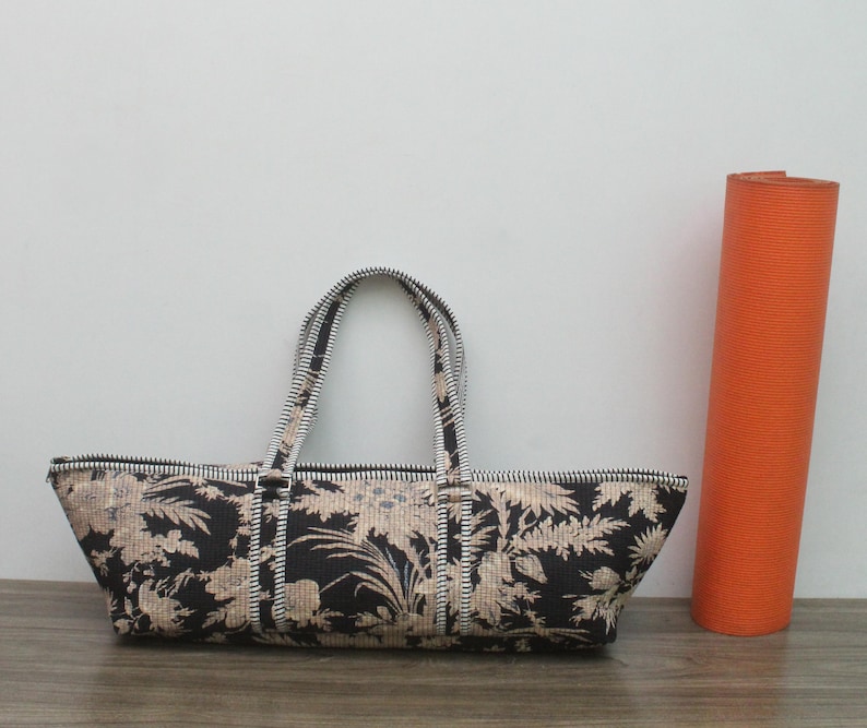 May include: A long, rectangular black and beige floral patterned bag with two matching handles and a top zipper closure. The bag features a detailed botanical print and is positioned next to a tall, solid orange cylindrical yoga mat.