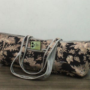 May include: A long, rectangular tote bag featuring a black fabric base with a tan floral pattern. The bag includes two striped handles and a small exterior pocket containing a light green smartphone. A zipper runs along the top edge of the bag.