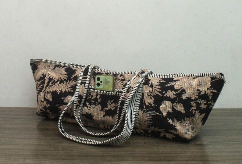 May include: A long, rectangular tote bag featuring a black fabric base with a tan floral pattern. The bag includes two striped handles and a small exterior pocket containing a light green smartphone. A zipper runs along the top edge of the bag.
