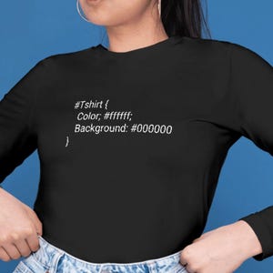 Pode incluir: Uma camisa preta de manga comprida com texto em código CSS branco que diz #Tshirt { Color: #ffffff; Background: #000000 }. A camisa tem gola redonda e é combinada com calças de ganga azul-claras com botão e fecho de correr visíveis.