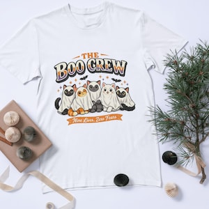 Op de afbeelding: Een wit t-shirt met korte mouwen en een afbeelding van vijf katten in spookkostuums. De tekst THE BOO CREW staat boven de katten en Nine Lives, Zero Fears op een banner eronder, omringd door kleine vleermuizen en sterren in oranje en zwart.