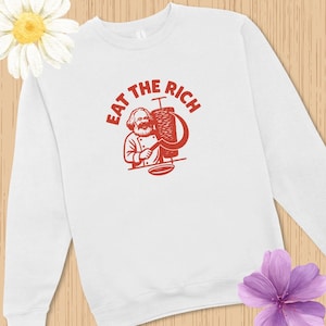 Puede incluir: Una sudadera blanca de manga larga con un estampado gráfico rojo de una figura barbuda junto a un asador vertical, acompañada de una hoz y el texto EAT THE RICH arqueado sobre la ilustración en una fuente roja llamativa.