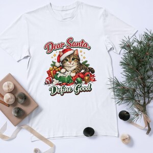 Könnte beinhalten: Ein weißes Kurzarm-T-Shirt mit einer Grafik eines braunen getigerten Katers, der eine rote Weihnachtsmannmütze trägt und von bunten Weihnachtsgeschenken umgeben ist. Der Text über der Katze lautet Dear Santa, darunter steht Define Good.