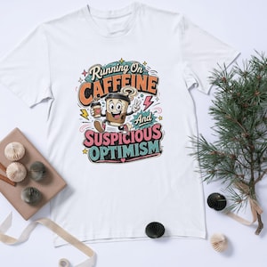 Op de afbeelding: Een wit T-shirt met korte mouwen en een kleurrijke afbeelding van een cartoon koffiebeker. De tekst luidt Running On CAFFEINE And SUSPICIOUS OPTIMISM. Het ontwerp bevat sterren, bliksemschichten en een hand die een vredesteken maakt.