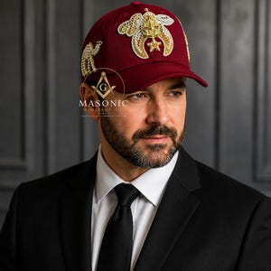May include: A deep burgundy baseball cap featuring intricate gold and white beaded embroidery on the front and side panels. The front displays a large ornate emblem, while the side features a camel motif. The text MASONIC HERITAGE CO is visible.
