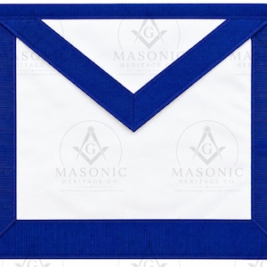 May include: A white Masonic apron featuring a prominent blue border and a triangular flap. The design includes a blue ribbon trim along the edges and the Masonic Heritage Co. logo with a square and compass symbol printed on the white fabric surface.