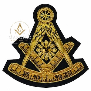 May include: A black fabric patch featuring gold bullion embroidery of a masonic square and compass symbol. The design includes a central sun face, a circular top emblem, and a graduated scale along the bottom edge with numerical markings.