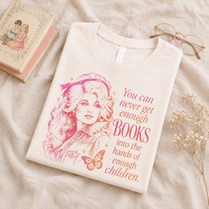 May include: A cream-colored t-shirt featuring a pink and orange graphic illustration of a woman with voluminous hair, a butterfly, and the text: You can never get enough BOOKS into the hands of enough children.