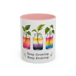 Puede incluir: Una taza de cerámica blanca con un borde interior rosa suave. El exterior presenta tres frascos de vidrio con esquejes de plantas verdes y raíces visibles. El agua en cada frasco muestra diferentes degradados de color. El texto dice: Keep Growing, Keep Evolving.