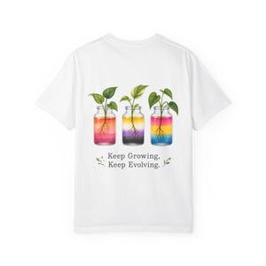 Puede incluir: Una camiseta blanca con un gráfico de tres frascos de vidrio, cada uno con una pequeña planta verde con raíces visibles. El líquido en cada frasco muestra diferentes degradados de color. Debajo de los frascos, el texto dice Keep Growing, Keep Evolving.