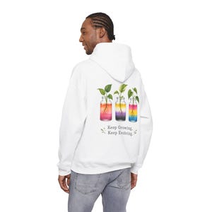 Puede incluir: Una sudadera con capucha blanca que presenta un gráfico en la parte posterior de tres frascos de vidrio con plantas y raíces. El líquido en los frascos muestra colores degradados y el texto debajo dice, Keep Growing, Keep Evolving.