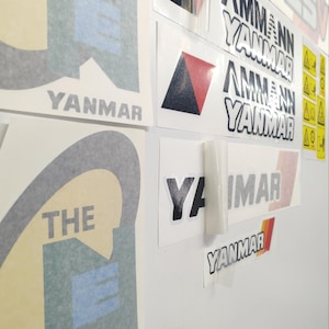 Op de afbeelding: Een verzameling zelfklevende stickers met de tekst AMMANN YANMAR en YANMAR in zwarte en grijze letters. Sommige stickers bevatten een rood en zwart driehoekig logo of een geel waarschuwingspictogram. De stickers staan op een witte ondergrond.