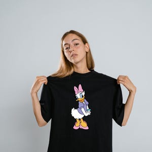 Może przedstawiać: Czarny t-shirt z kr&oacute;tkim rękawem z nadrukiem graficznym postaci z kresk&oacute;wki Daisy Duck. Postać przedstawiona jest z białymi pi&oacute;rami, w fioletowej koszulce, r&oacute;żowej kokardzie i r&oacute;żowych butach, stojąca w klasycznej pozie na przodzie koszulki.