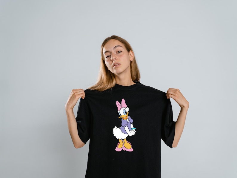 Może przedstawiać: Czarny t-shirt z kr&oacute;tkim rękawem z nadrukiem graficznym postaci z kresk&oacute;wki Daisy Duck. Postać przedstawiona jest z białymi pi&oacute;rami, w fioletowej koszulce, r&oacute;żowej kokardzie i r&oacute;żowych butach, stojąca w klasycznej pozie na przodzie koszulki.