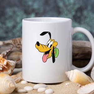 Może przedstawiać: Biały ceramiczny kubek z grafiką przedstawiającą postać z kresk&oacute;wki Pluto. Pies ma ż&oacute;łte futro, czarne uszy i r&oacute;żowy język, a jego wyraz pyska jest radosny. Kubek ma standardowy cylindryczny kształt oraz wygodny uchwyt do trzymania.