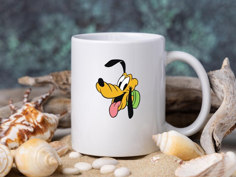 Może przedstawiać: Biały ceramiczny kubek z grafiką przedstawiającą postać z kresk&oacute;wki Pluto. Pies ma ż&oacute;łte futro, czarne uszy i r&oacute;żowy język, a jego wyraz pyska jest radosny. Kubek ma standardowy cylindryczny kształt oraz wygodny uchwyt do trzymania.