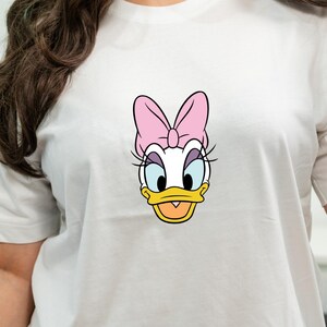 Może przedstawiać: Biała koszulka z kr&oacute;tkim rękawem z centralną grafiką postaci z kresk&oacute;wki Daisy Duck. Postać ma białe pi&oacute;ra, ż&oacute;łty dzi&oacute;b, niebieskie powieki, długie czarne rzęsy i dużą r&oacute;żową kokardę umieszczoną na czubku głowy.