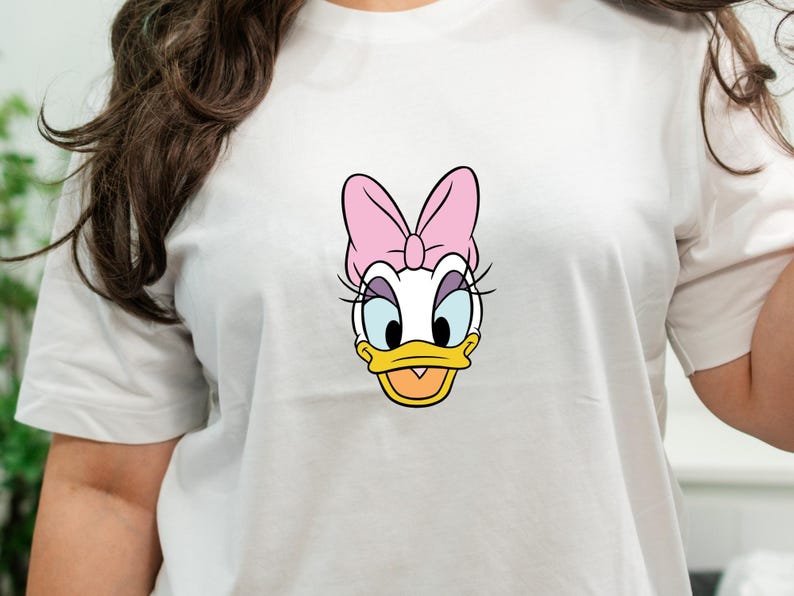 Może przedstawiać: Biała koszulka z kr&oacute;tkim rękawem z centralną grafiką postaci z kresk&oacute;wki Daisy Duck. Postać ma białe pi&oacute;ra, ż&oacute;łty dzi&oacute;b, niebieskie powieki, długie czarne rzęsy i dużą r&oacute;żową kokardę umieszczoną na czubku głowy.