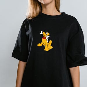 Może przedstawiać: Czarny t-shirt z kr&oacute;tkim rękawem z centralną grafiką przedstawiającą ż&oacute;łtego psa z kresk&oacute;wki Pluto, kt&oacute;ry ma na sobie czerwoną obrożę i trzyma w pysku białą kość. Koszulka ma okrągły dekolt oraz luźny kr&oacute;j z szerokimi rękawami.