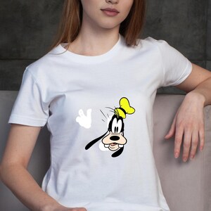 Może przedstawiać: Biały t-shirt z kr&oacute;tkim rękawem z nadrukiem graficznym przedstawiającym postać z kresk&oacute;wki Goofy'ego. Postać jest ukazana w ż&oacute;łtym kapeluszu, z czarnymi uszami i białą rękawicą uniesioną w geście powitania na środku koszulki.
