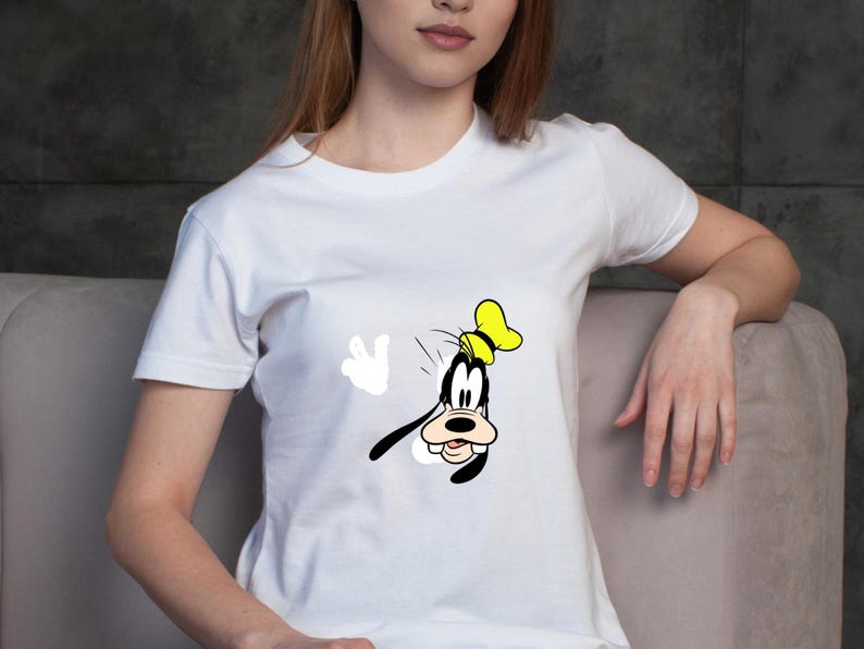 Może przedstawiać: Biały t-shirt z kr&oacute;tkim rękawem z nadrukiem graficznym przedstawiającym postać z kresk&oacute;wki Goofy'ego. Postać jest ukazana w ż&oacute;łtym kapeluszu, z czarnymi uszami i białą rękawicą uniesioną w geście powitania na środku koszulki.