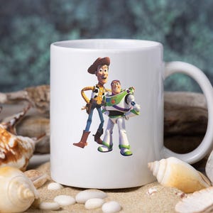 May include: A white ceramic mug featuring a colorful graphic of the characters Woody and Buzz Lightyear from the Toy Story film franchise. The mug has a standard cylindrical shape with a side handle and a smooth, glossy finish on the exterior.