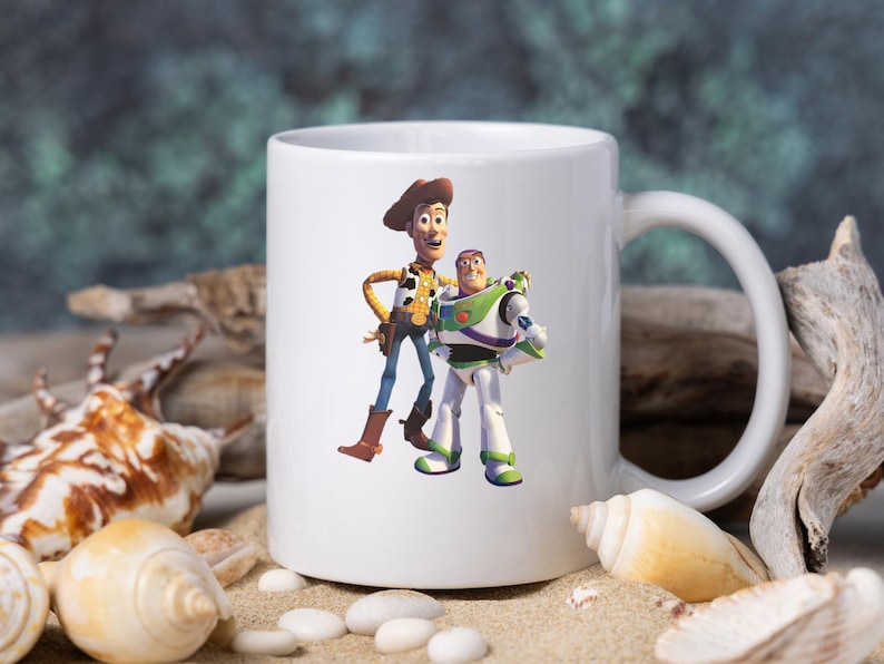 May include: A white ceramic mug featuring a colorful graphic of the characters Woody and Buzz Lightyear from the Toy Story film franchise. The mug has a standard cylindrical shape with a side handle and a smooth, glossy finish on the exterior.