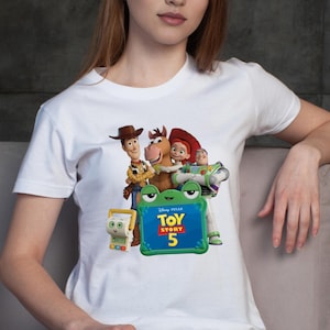 May include: A white short-sleeved t-shirt featuring a graphic print of characters from Toy Story including Woody, Bullseye, Jessie, and Buzz Lightyear, along with a green frog-shaped frame containing the text Disney Pixar Toy Story 5.