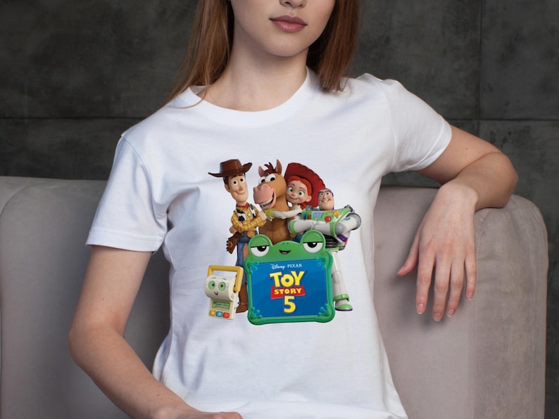 May include: A white short-sleeved t-shirt featuring a graphic print of characters from Toy Story including Woody, Bullseye, Jessie, and Buzz Lightyear, along with a green frog-shaped frame containing the text Disney Pixar Toy Story 5.