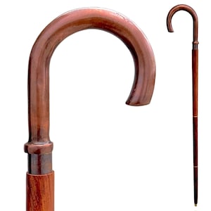 Pode incluir: Uma bengala com um cabo curvo em tom de cobre polido e um eixo de madeira castanho-escuro. O cabo liga-se ao eixo com um colar metálico e a peça inteira apresenta um acabamento liso com padrões de grão de madeira visíveis.