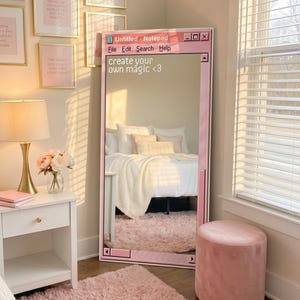 Pode incluir: Um espelho de chão retangular alto com uma moldura rosa em design de janela de computador com o texto 'Untitled - Notepad', 'File Edit Search Help' e 'create your own magic <3'. O espelho reflete uma cama com roupa de cama branca.