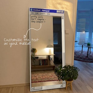 Pode incluir: Um espelho retangular de corpo inteiro com uma moldura branca impressa com uma interface clássica de bloco de notas de computador. O vidro exibe o texto 'pretty girls don't compete - they inspire ♡' e uma seta apontando para o texto com a legenda 'Customize the text on your mirror!'