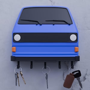 Pode incluir: Um porta-chaves de parede azul com o formato da frente de uma carrinha. Possui um para-brisas preto com limpa para-brisas, dois faróis circulares brancos e dois pequenos piscas cor de laranja. Cinco ganchos pretos seguram três conjuntos de chaves.