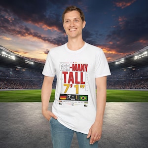 Pode incluir: Uma t-shirt branca de manga curta com um gráfico que inclui o texto GER-MANY TALL 7'1" e um placar mostrando um resultado de 7-1 entre as bandeiras da Alemanha e do Brasil. O design inclui uma bola de futebol, um ícone de torre e texto.
