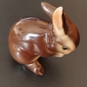 Op de afbeelding: Een klein, glanzend keramieken beeldje van een konijn in zittende houding. Het konijn heeft een donkerbruin lichaam met lichtere beige accenten op het gezicht en de oren, met een gladde, reflecterende afwerking en gedetailleerde gelaatstrekken.