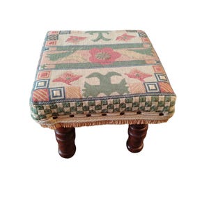 May include: A square footstool featuring a woven fabric top with a multi-coloured floral and geometric pattern in shades of pink, green, and beige. The base is trimmed with decorative metal nailheads and fringe, supported by four dark brown wooden legs.
