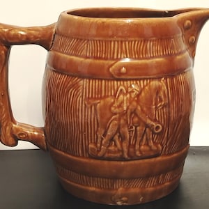 Op de afbeelding: Een bruin geglazuurde keramische kan in de vorm van een houten vat met een handvat dat eruitziet als een boomtak. De buitenkant heeft een reliëf van een persoon die naast een paard staat, met horizontale banden rondom het lichaam.