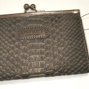 May include: A grey rectangular purse with a snakeskin-like embossed texture and a silver-toned metal kiss-lock clasp. The front features a rectangular leather patch embossed with the text OSPREY LONDON. A thin black string tag is attached to the side.