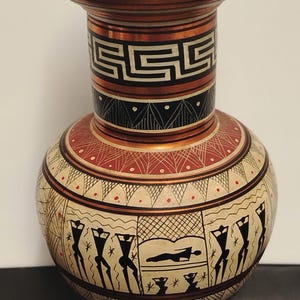 Könnte beinhalten: Eine dekorative Vase mit kupferfarbenem Rand und Fuß sowie einem cremefarbenen Körper. Der Hals zeigt ein schwarz-weißes Mäandermuster. Der Körper ist mit schwarzen Silhouettenfiguren, geometrischen Linien und roten Punkten verziert.