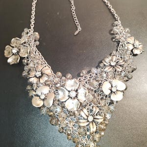Può includere: Una collana importante color argento caratterizzata da molteplici ciondoli a forma di fiore in metallo con cristalli trasparenti al centro, intervallati da grappoli di perline grigie sfaccettate. Il design è unito da una catena argentata con chiusura a moschettone.