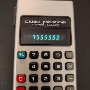 May include: A silver Casio pocket-mini electronic calculator with a black display screen showing the number 7555999 in green digits. The device features a grid of black, red, blue, and grey buttons with white numerals and mathematical symbols.