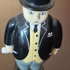 Op de afbeelding: Een plastic figuurtje van een gezet personage met een zwarte hoge hoed, een zwart jasje met witte rand, een lichtgeel vest met zwarte knopen, een wit overhemd met een zwarte stropdas en een grijze broek, staand op een zwarte voet met opwindsleutel.