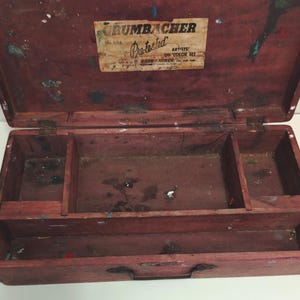 Pode incluir: Uma caixa de madeira vintage Grumbacher Pre-tested Artists' Oil Color Set com tampa articulada. O interior possui vários compartimentos para armazenamento, mostrando sinais de uso com vários respingos de tinta na superfície de madeira.