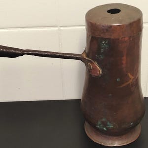 May include: A tall, cylindrical copper vessel with a dark, weathered patina and visible green oxidation. It features a long, dark, horizontal metal handle attached to the side and a circular opening at the top, resting on a flared base.